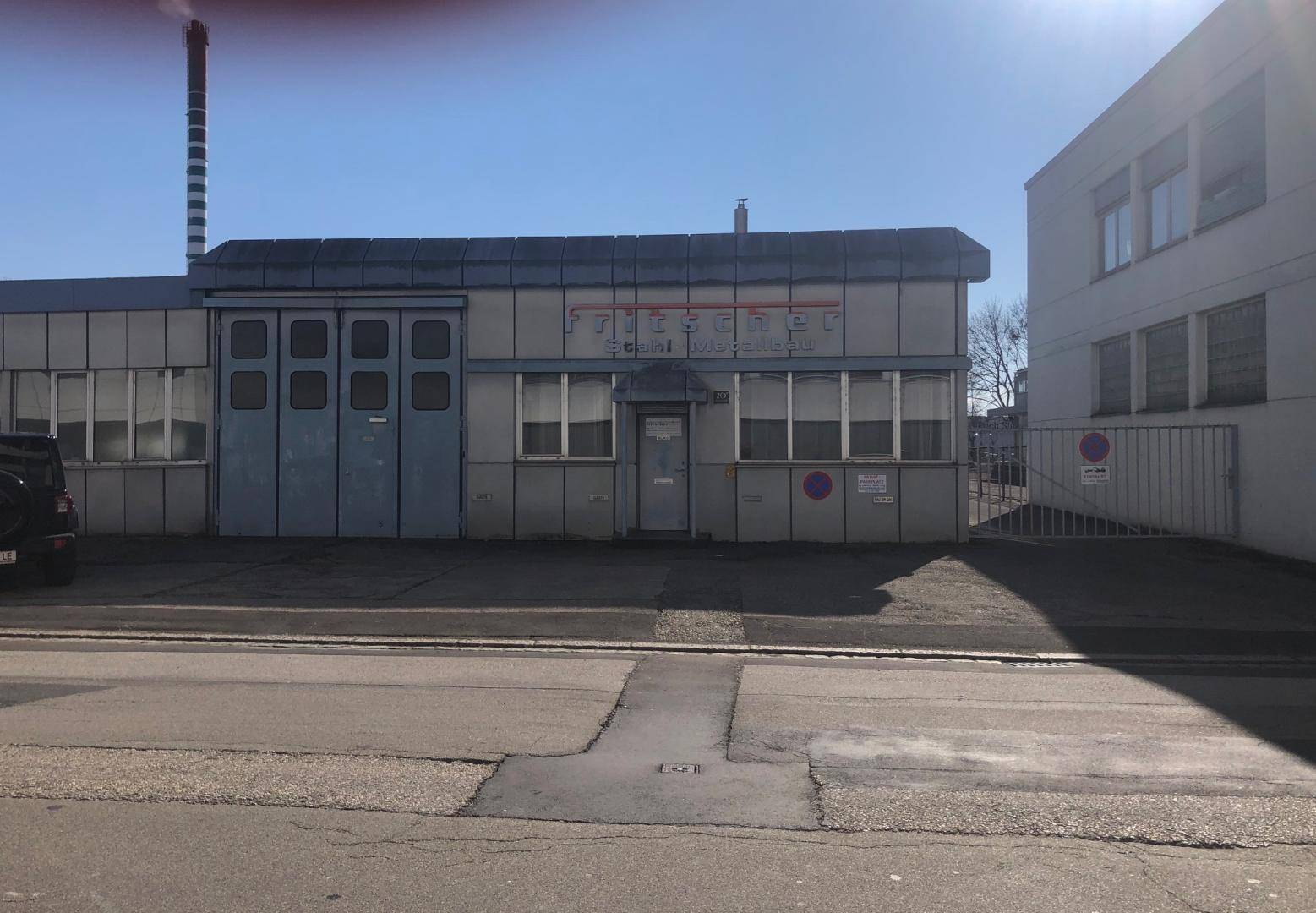 Ansicht der Gewerbeimmobilie von der Straße mit einem großen Schornstein im Hintergrund und Parkmöglichkeiten davor.