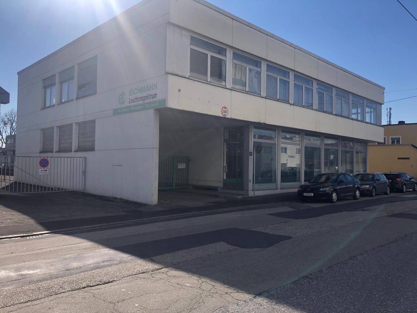 Mehrstöckiges Geschäftsgebäude mit weißer Fassade, großen Fensterfronten und Parkplätzen an der Straße in der Sonne.