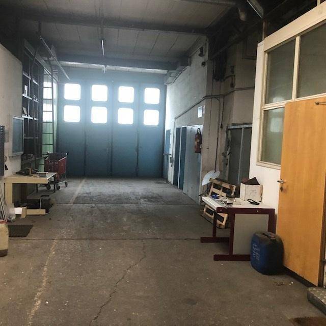 Werkstattbereich mit Betonboden, einem großen Rolltor mit Fenstern und einer Holztür, bereit für verschiedene Arbeiten.