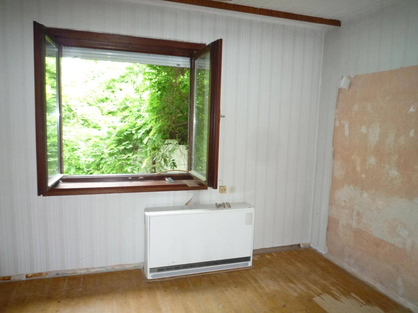 Leerer Raum mit Holzdielenboden, Heizkörper und Fenster mit Blick ins Grüne.