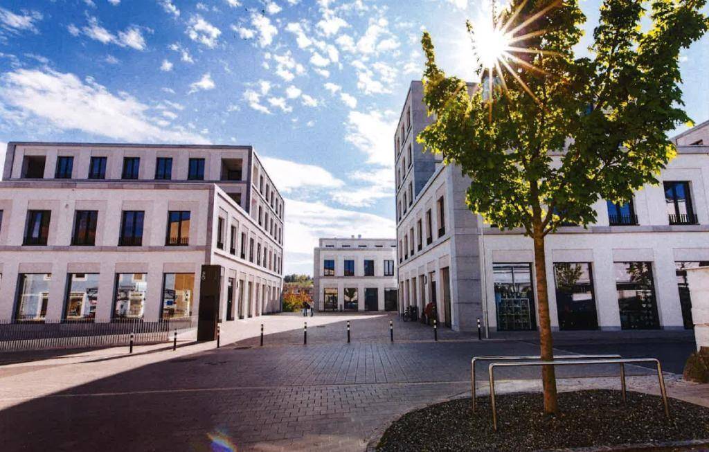 Moderne Gebäude umgeben einen gepflasterten Platz, beleuchtet von der Sonne unter blauem Himmel.