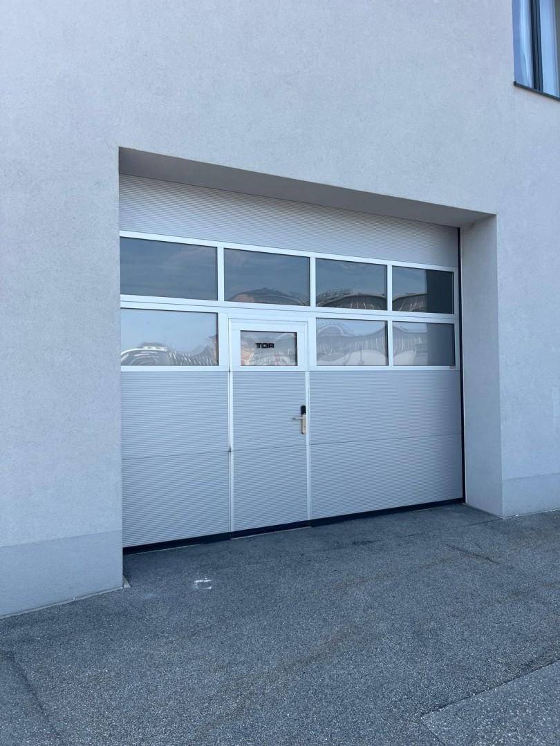 Großes Garagentor mit Fenstern in einer hellen Gebäudefassade, bietet Zugang zu einer Lagerhalle.