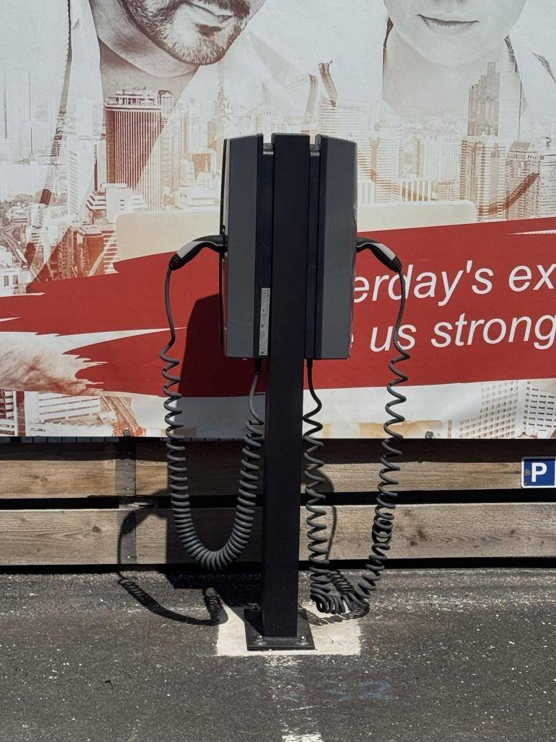Ladestation für Elektrofahrzeuge mit zwei Ladekabeln, vor einer großen Werbetafel auf dem Parkplatz.
