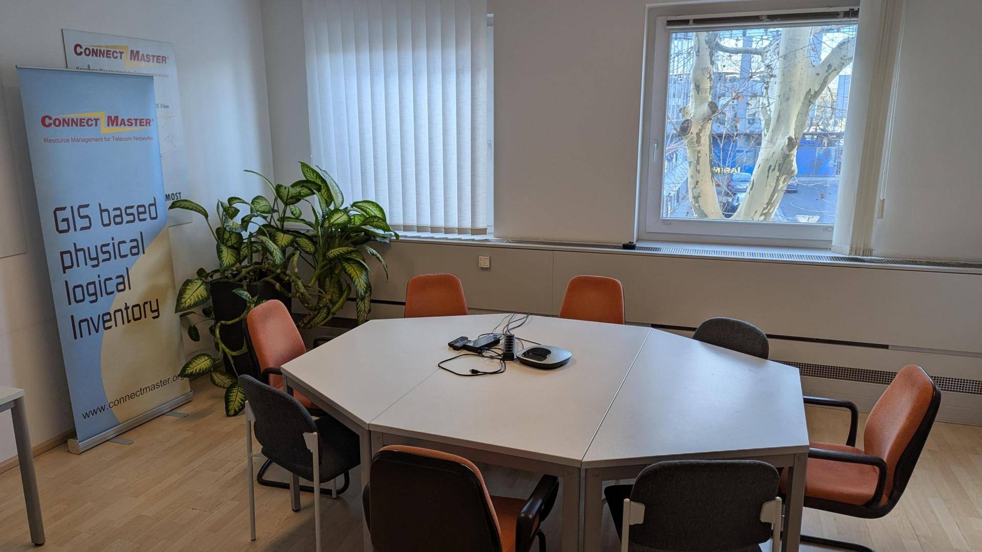 Konferenzraum mit einem großen achteckigen Tisch, mehreren Stühlen und einem Fenster mit Jalousien.