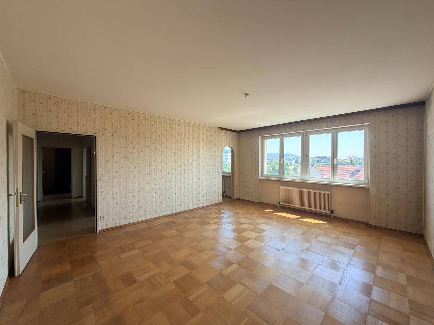 Helles Wohnzimmer mit Parkettboden, gemusterter Tapete und großen Fenstern mit Weitblick.