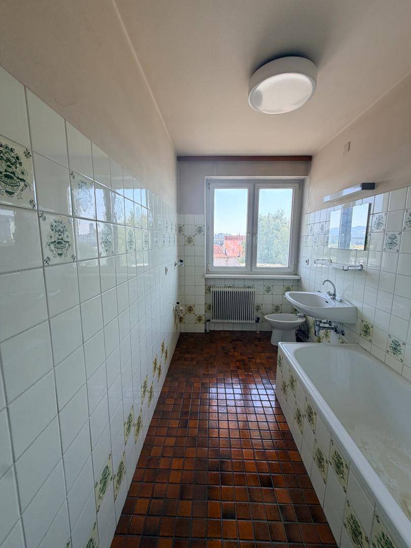 Badezimmer mit Badewanne, Waschbecken, Bidet und Fenster, gefliest im 80er-Jahre-Stil.
