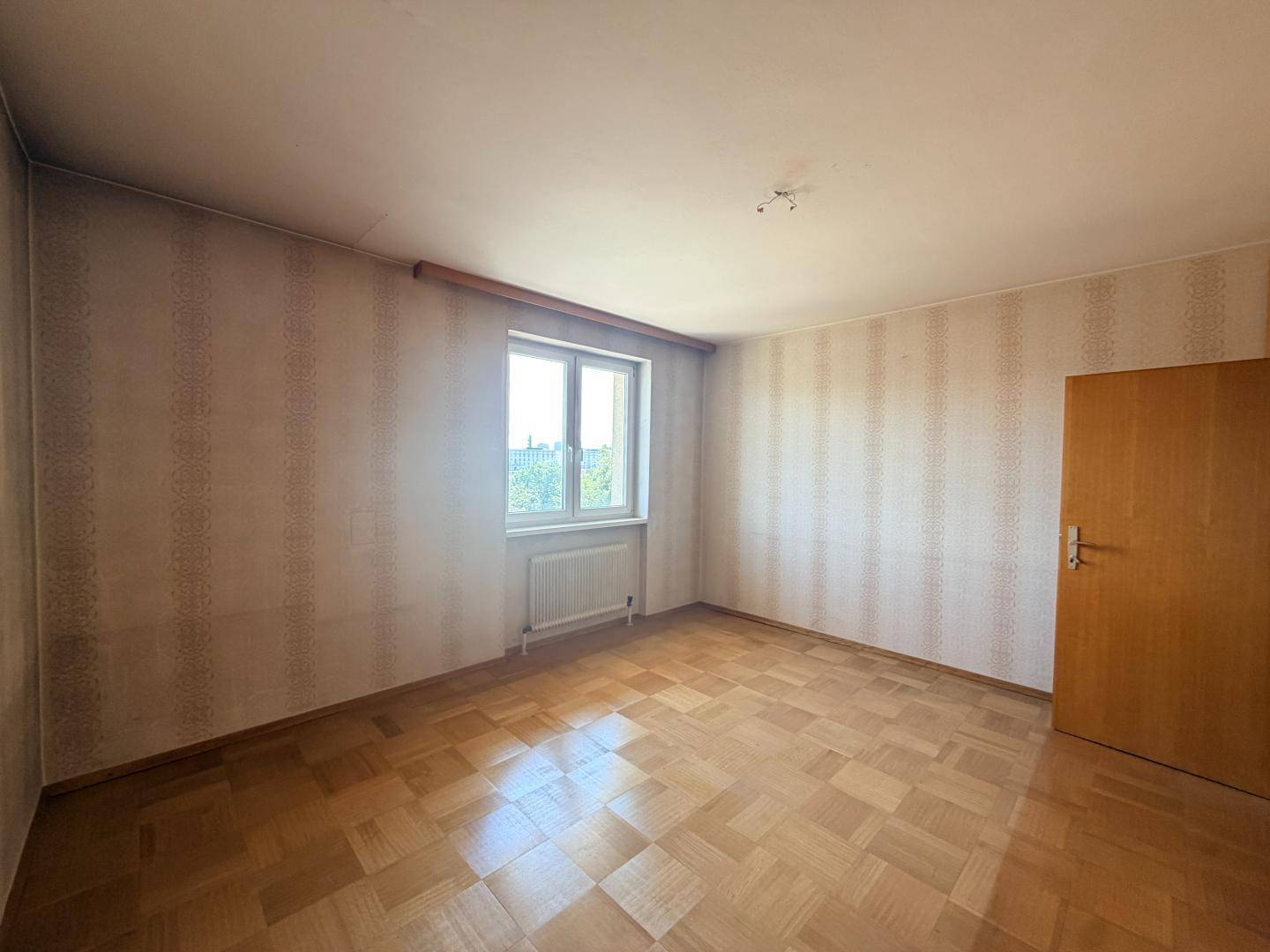 Leeres Zimmer mit Parkettboden, gemusterter Tapete und einem Fenster.