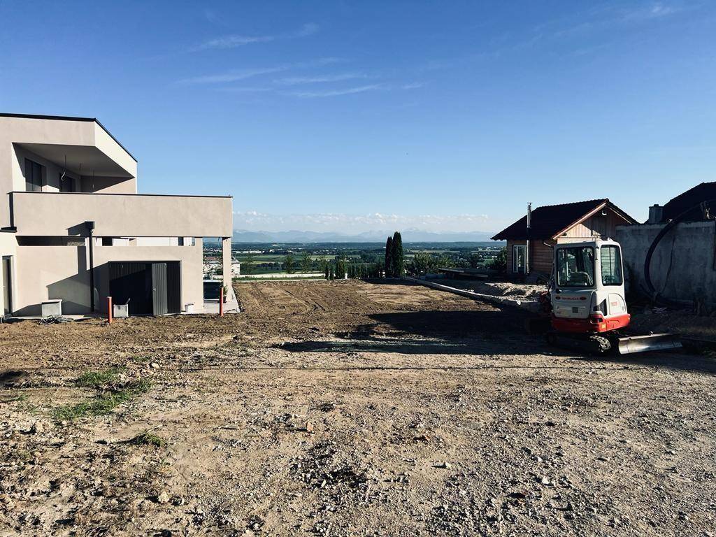 Blick über ein unbebautes Grundstück mit moderner Architektur und weitem Landschaftsblick bis zu den Bergen.