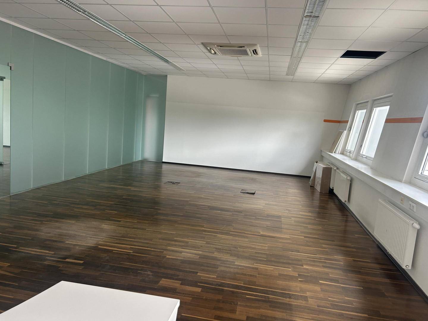 Geräumiges Büro mit dunklem Parkettboden, hellen Wänden und abgehängter Decke.