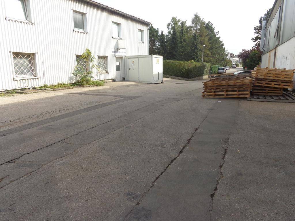 Außenbereich eines Industriegebäudes mit gepflastertem Hof, gestapelten Paletten und einem Container.