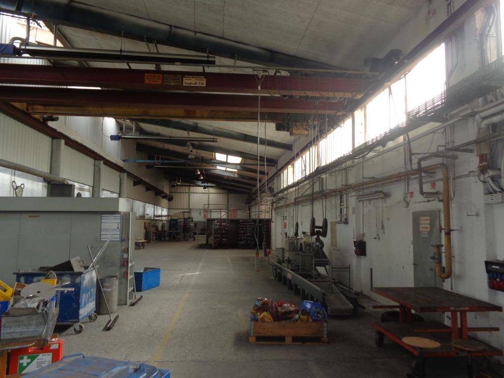 Lange Industriehalle mit hohen Decken, Oberlichtern und einem Kran, gefüllt mit Arbeitsmaterialien.