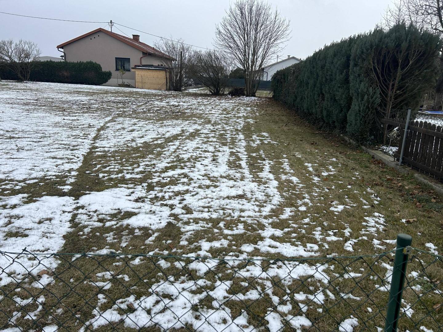 Grundstück mit Rasenfläche und Schneeresten, umgeben von einem Zaun und Häusern.