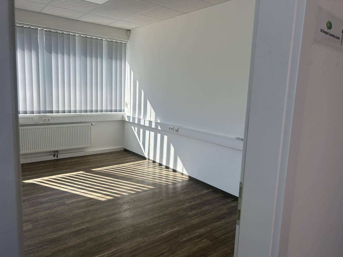 Leerer Büroraum mit Holzfußboden, großen Fenstern und Jalousien, gut beleuchtet durch natürliches Licht.