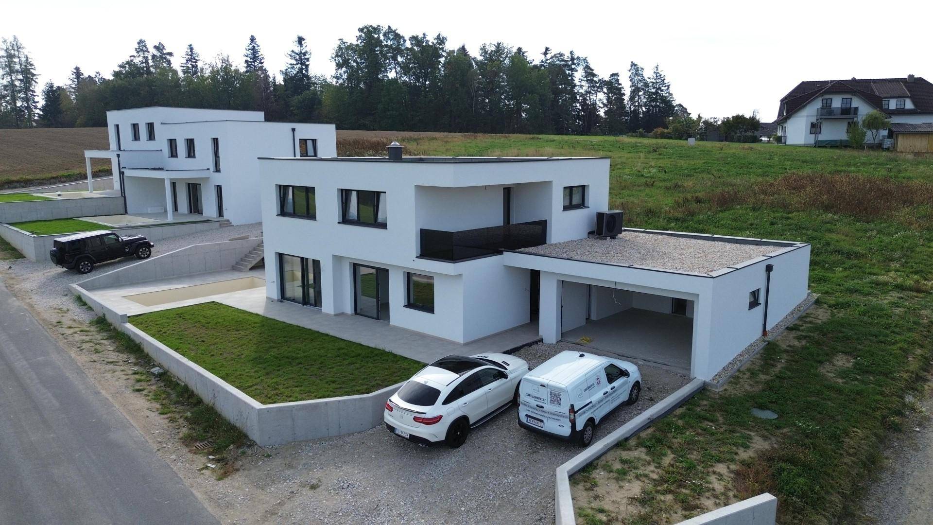 Zwei moderne weiße Häuser mit Flachdächern und Carports, umgeben von grünen Feldern und Bäumen.