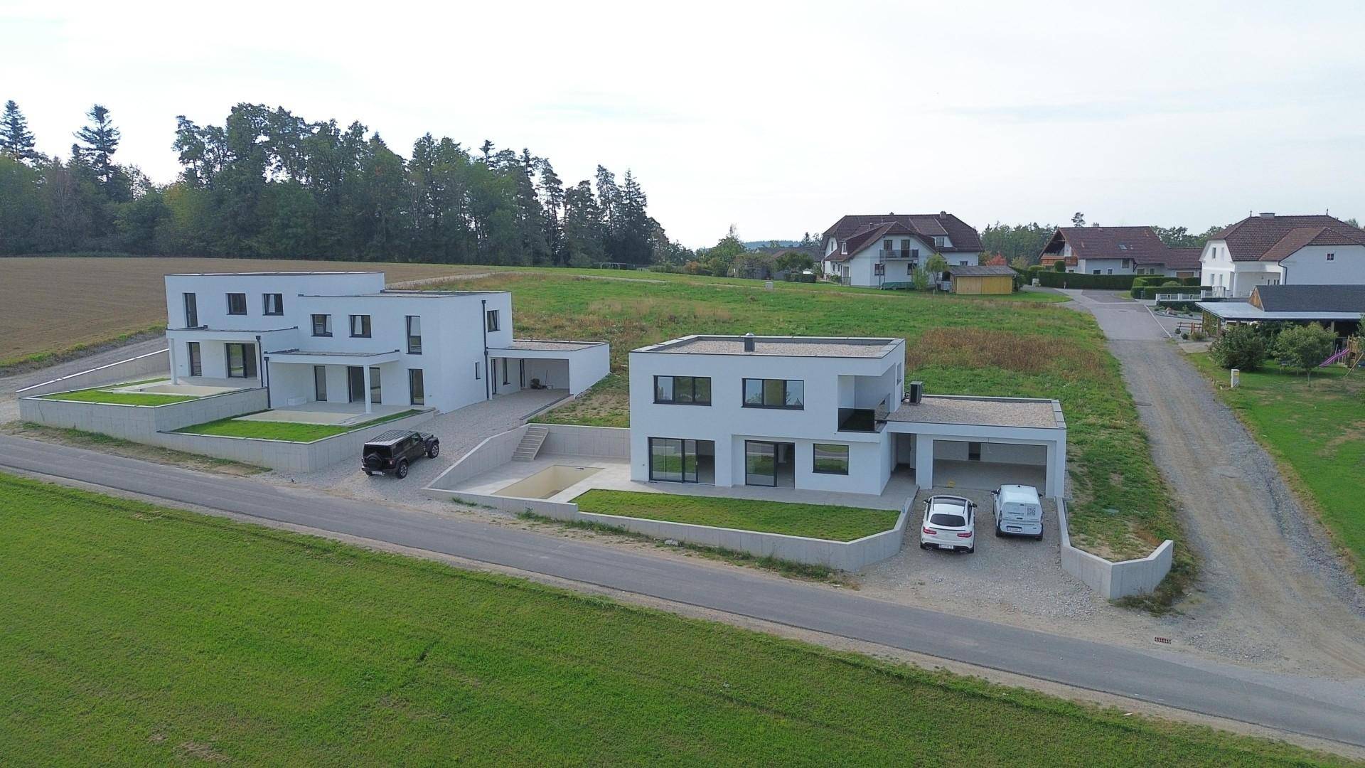 Luftaufnahme von zwei modernen weißen Häusern mit Flachdächern, eingebettet in eine grüne Landschaft.