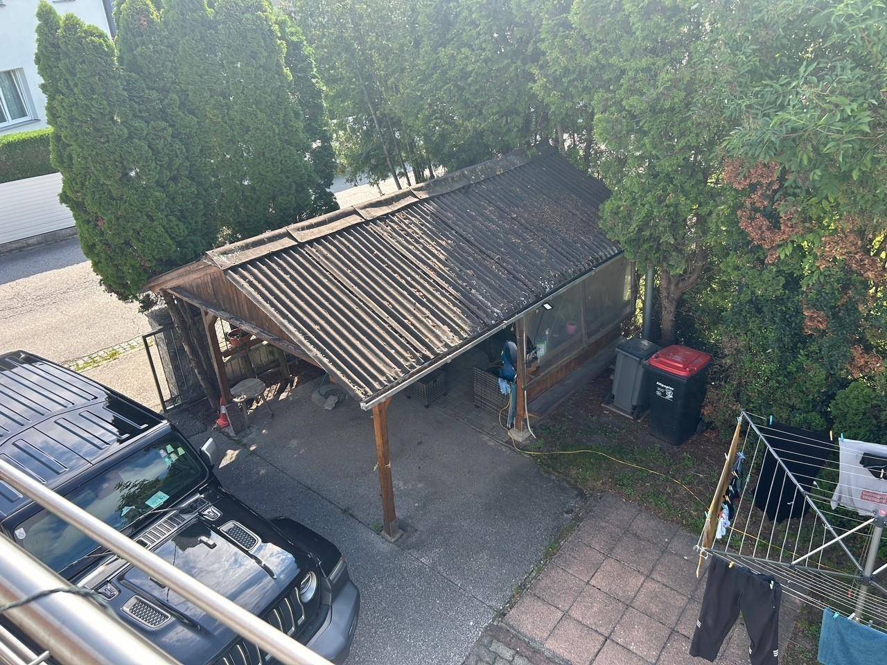 Überdachter Carport-Bereich mit Holzbalken und Wellblechdach, daneben ein Wäscheständer.