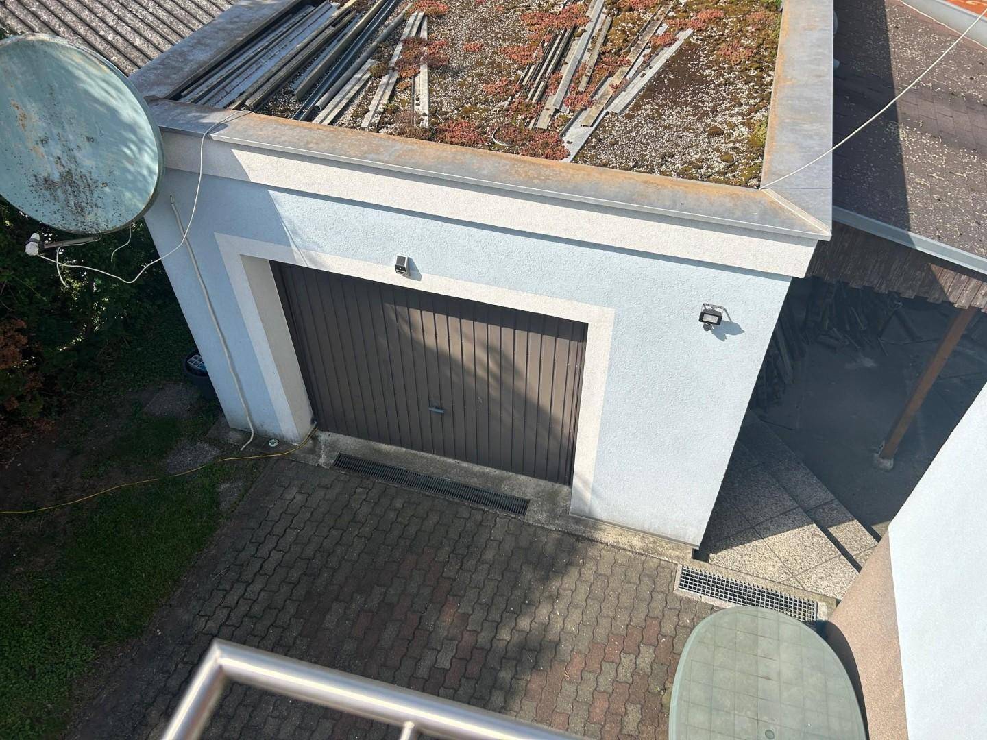 Einzelgarage mit braunem Tor und gepflasterter Zufahrt, daneben ein Satellitenspiegel.