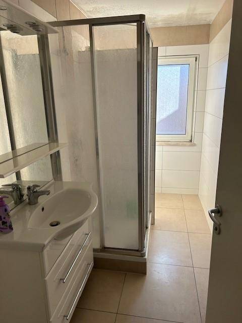 Funktionales Badezimmer mit Duschkabine, Einzelwaschbecken und einem Fenster, hell gefliest.