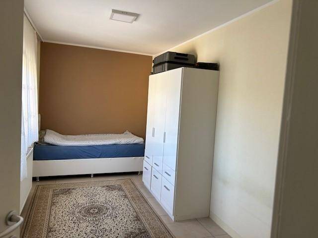 Schlafzimmer mit Einzelbett, weißem Kleiderschrank und einem gemusterten Teppich auf hellem Fliesenboden.