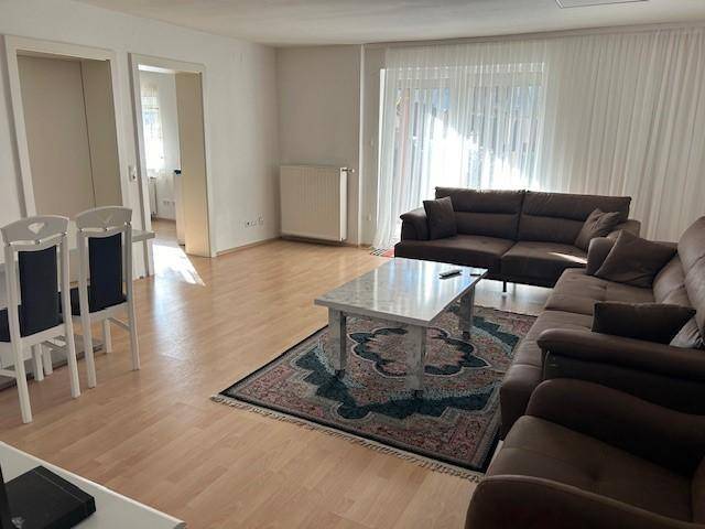 Heller Wohn- und Essbereich mit großem Ecksofa, Holzboden und Zugang zu einem Fenster mit Vorhängen.