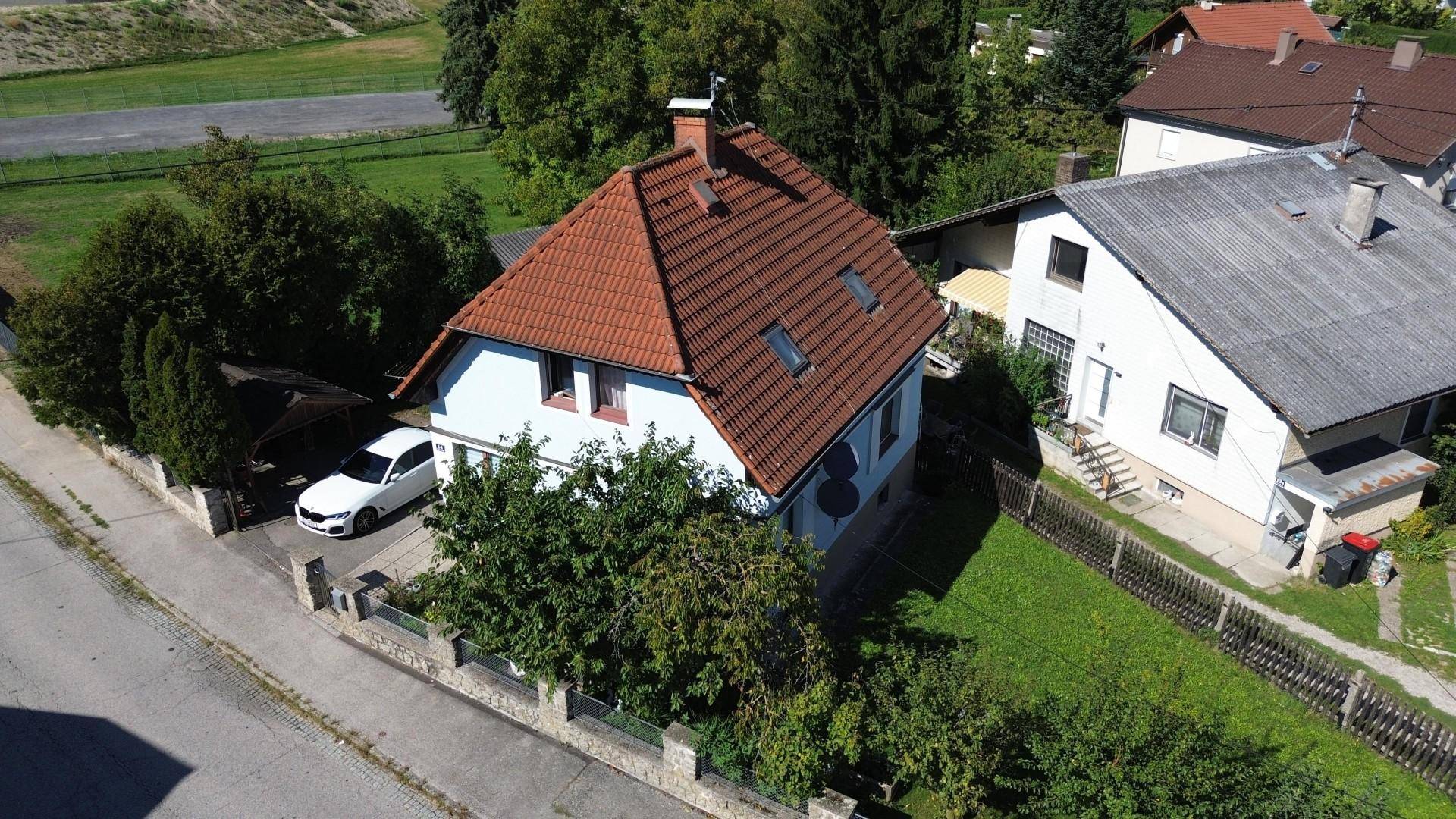 Vogelperspektive eines Wohnhauses mit großem Garten und angrenzender Straße unter blauem Himmel.
