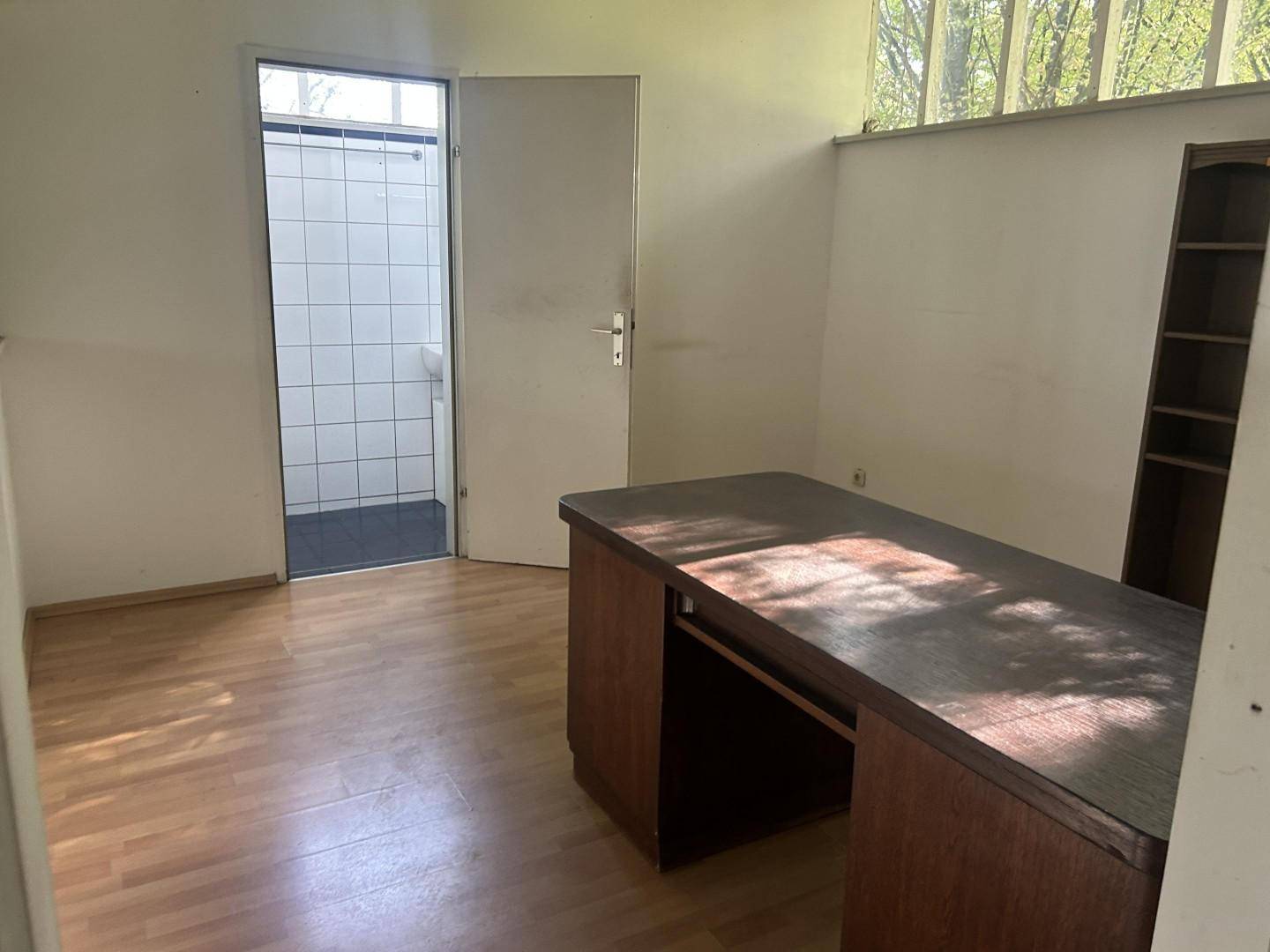 Kleines Büro mit Holzboden, einem Schreibtisch und Blick auf einen gefliesten Nebenraum mit Fenster.