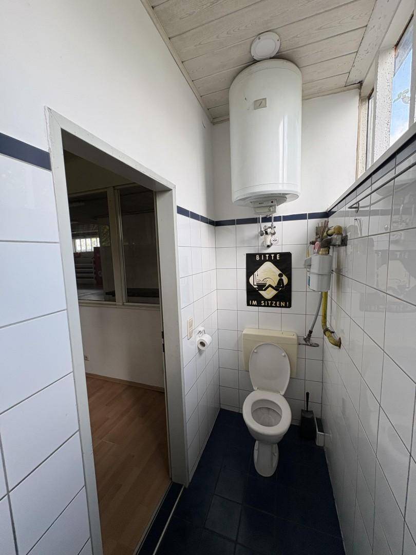 Kleine Toilette mit weißen Fliesen, blauem Streifen und einem Warmwasserboiler über der Toilette.