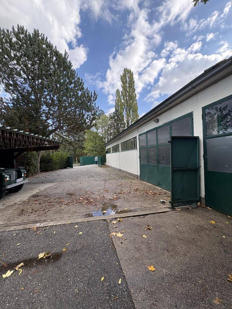 Außenansicht einer Gewerbeimmobilie mit Zufahrt, Parkplatz und angrenzendem Gebäude unter blauem Himmel.