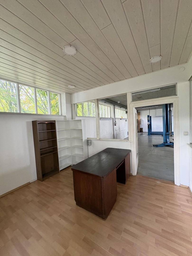 Büro mit Holzboden, Schreibtisch und Blick durch Glaswände in die angrenzende Werkstatt.