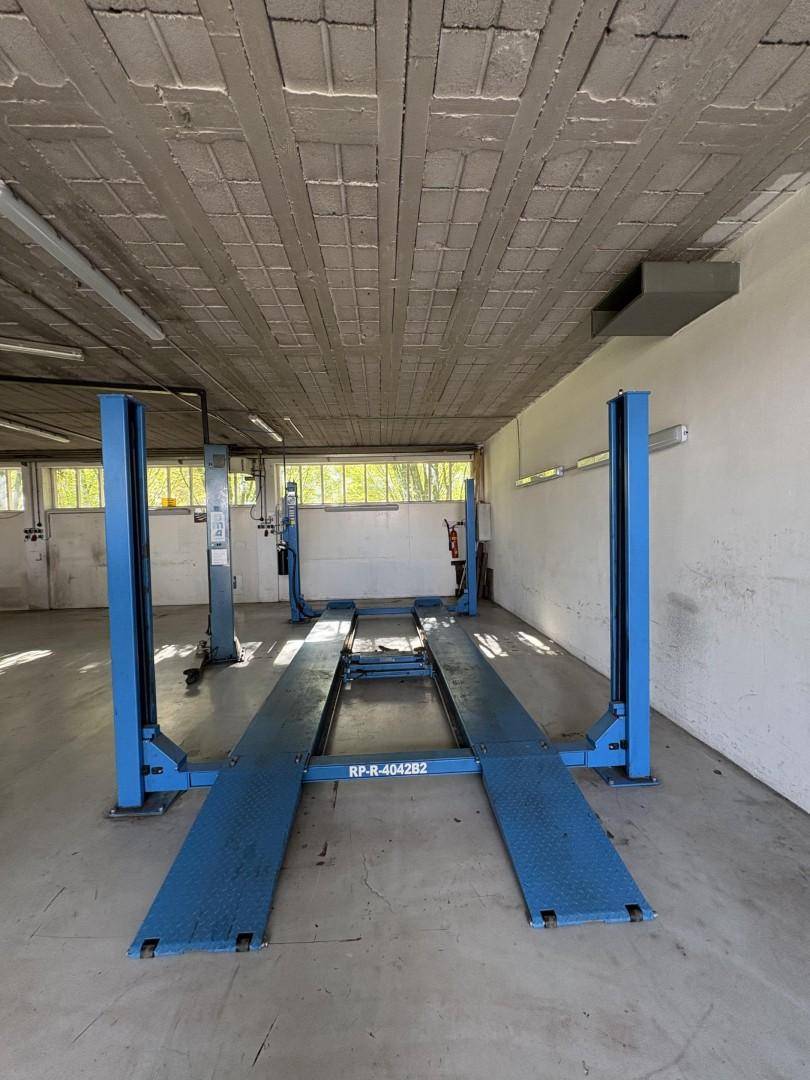 Werkstattbereich mit einer Vier-Säulen-Hebebühne und Betonboden, gut beleuchtet durch Fenster.