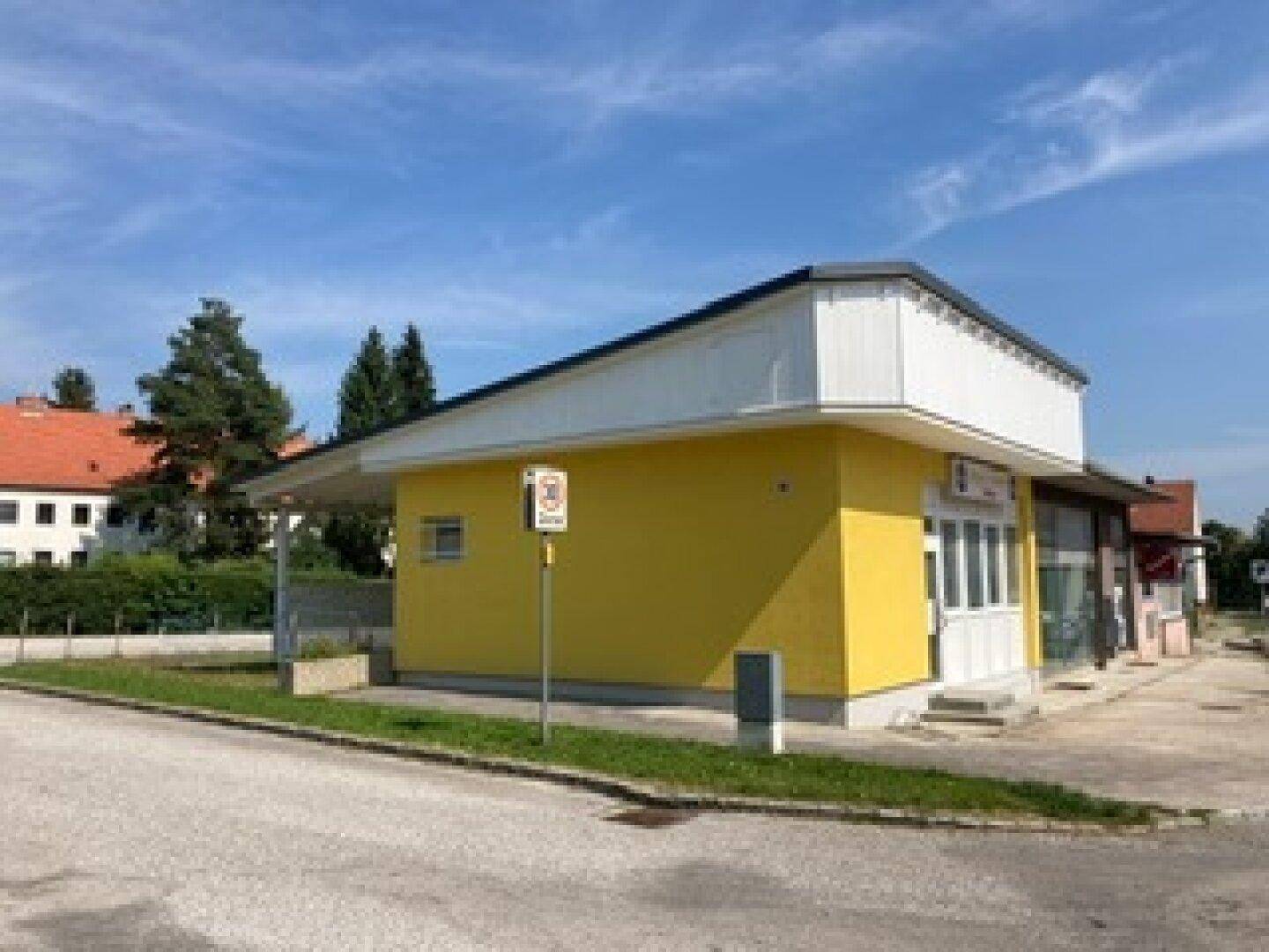 Gelbes Gebäude mit weißem Dach und Vordach, umgeben von einer Straße und Grünflächen unter blauem Himmel.