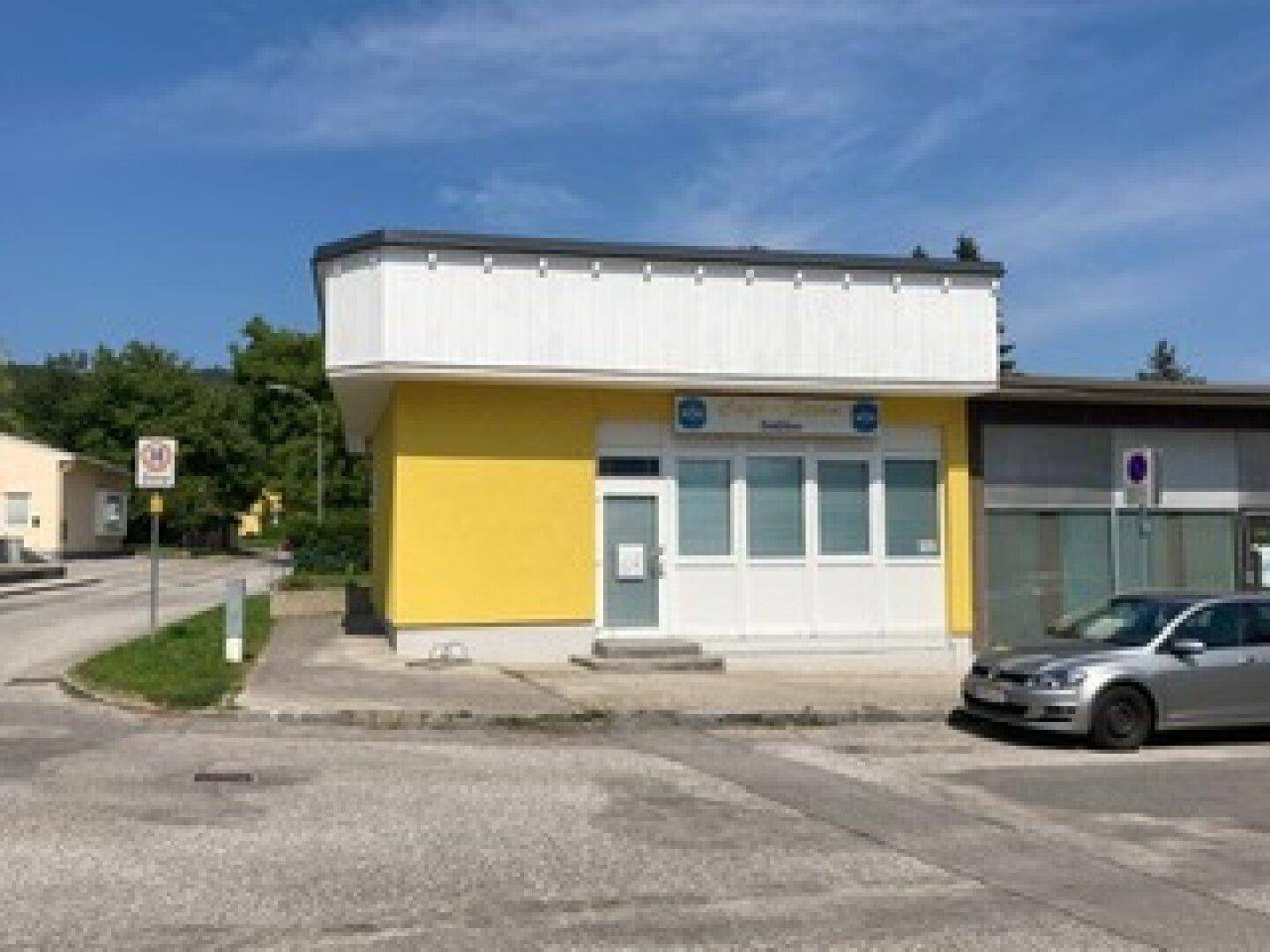 Gelbes Gebäude mit weißer Fassade und großen Fenstern, davor ein geparktes Auto auf dem Parkplatz.