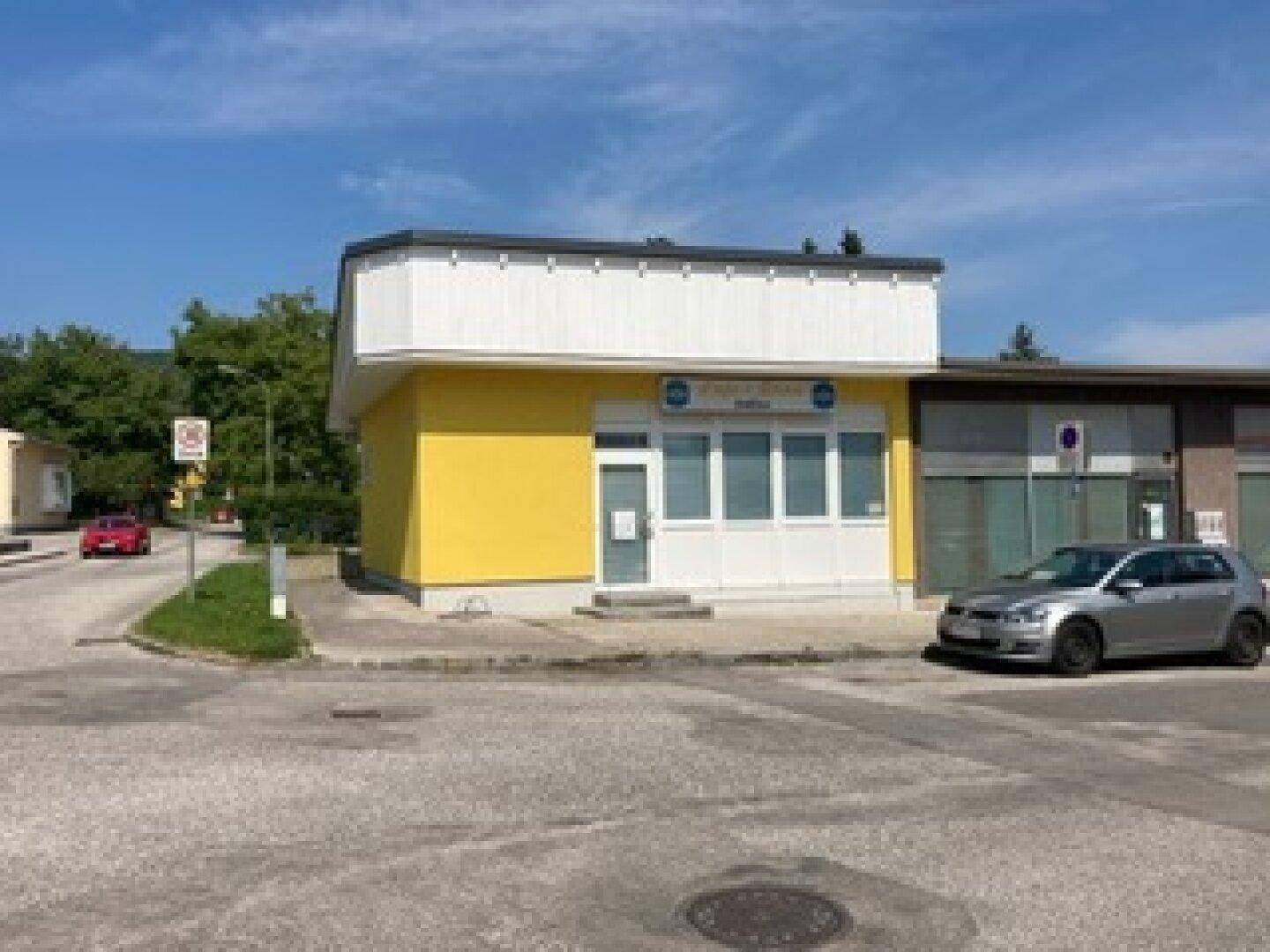 Gelbes Gebäude mit weißer Fassade und großen Fenstern, davor ein geparktes Auto auf dem Parkplatz.