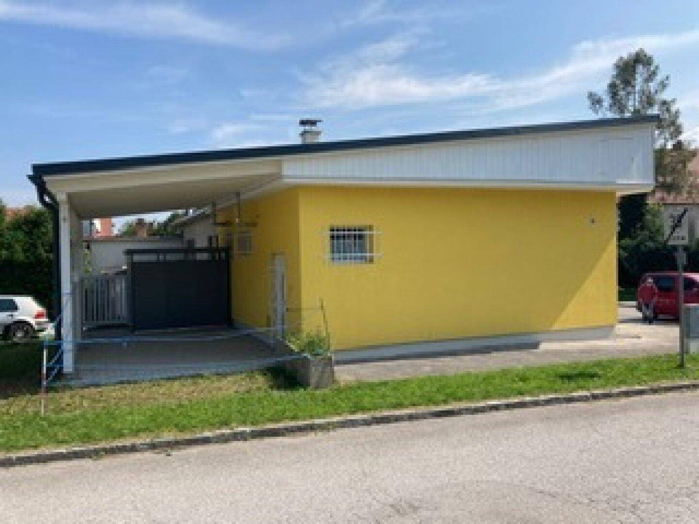 Gelbes Gebäude mit modernem Carport und gepflegter Rasenfläche an der Straße.
