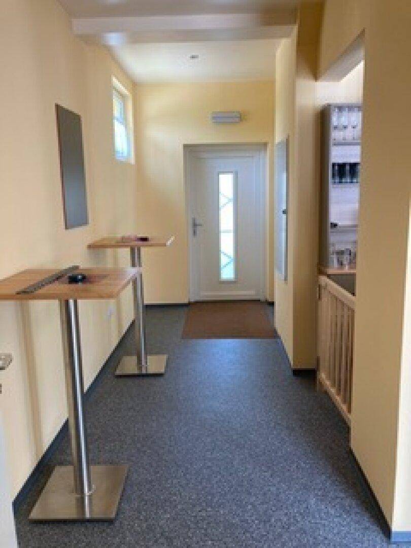 Heller Eingangsbereich mit zwei Stehtischen, dunklem Boden und einer weißen Tür am Ende des Flurs.