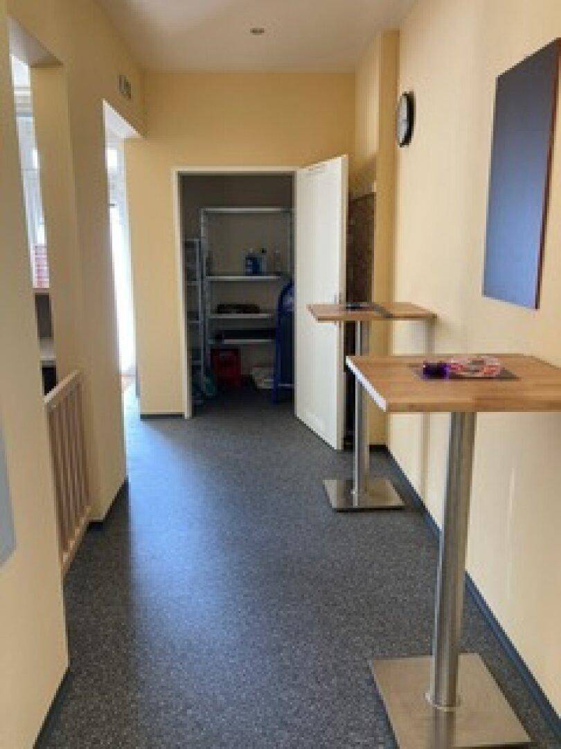 Heller Flur mit Stehtischen und einem offenen Abstellraum mit Regalen im Hintergrund.