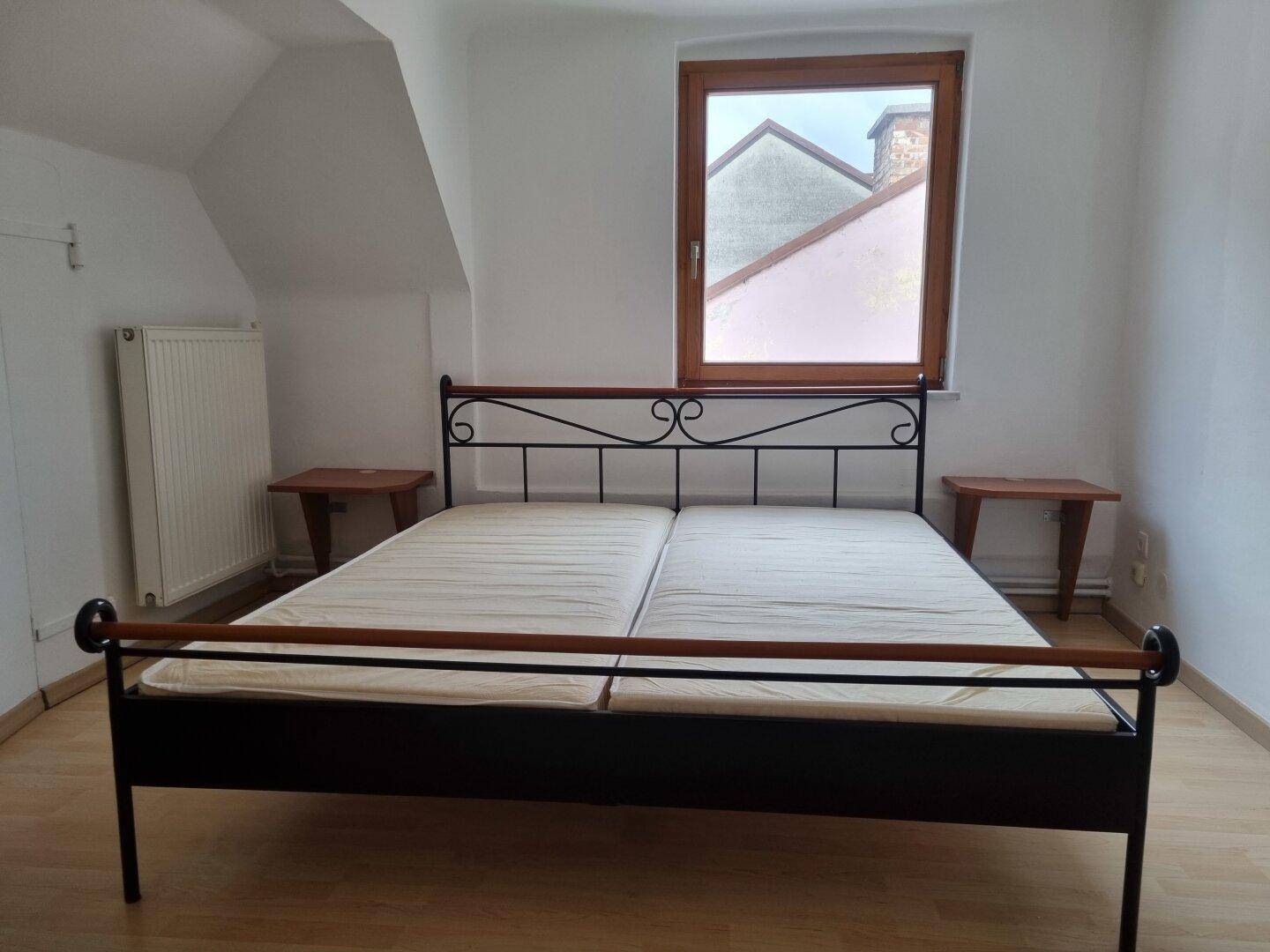 Schlafzimmer mit Metallbett, Holzboden, Fenster und Heizkörper unter der Dachschräge.