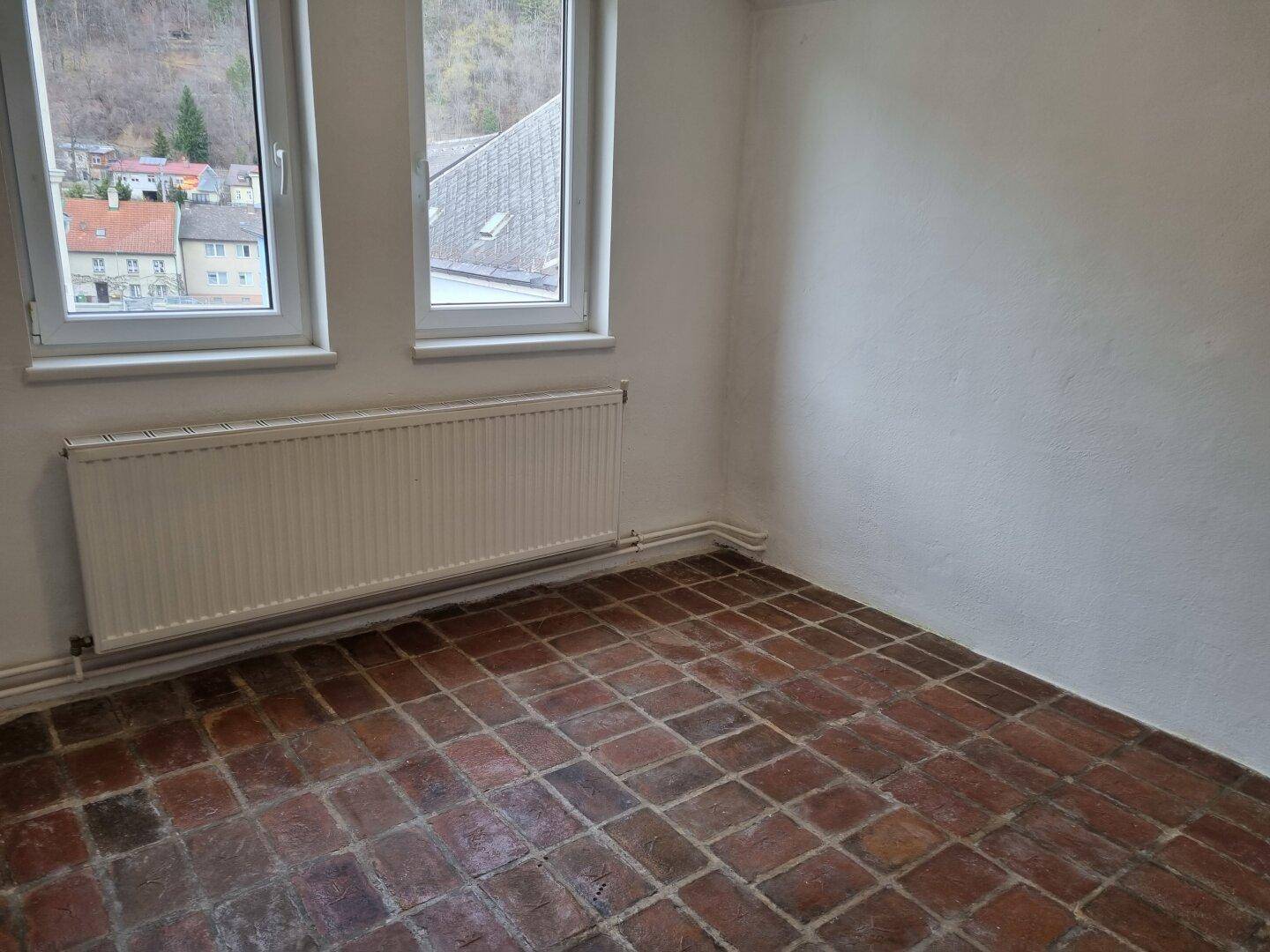 Heller Raum mit Terrakotta-Fliesenboden, Heizkörper und Fenstern mit Blick ins Grüne.
