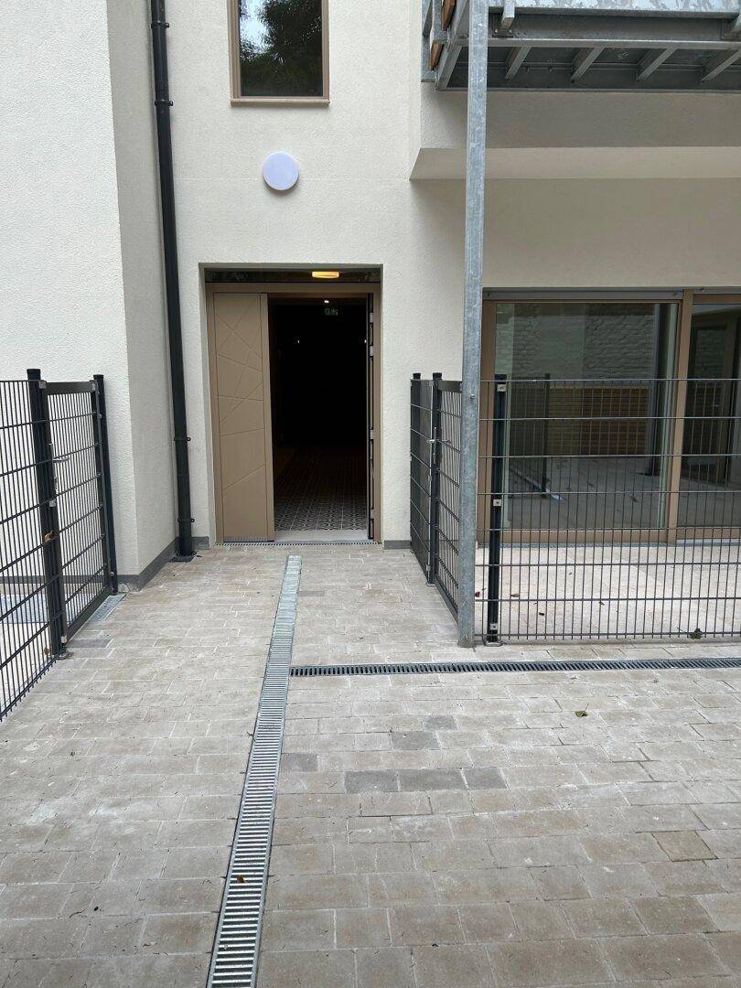 Eingangsbereich eines Neubaus mit gepflastertem Vorplatz, Metallzaun und moderner Fassadengestaltung.