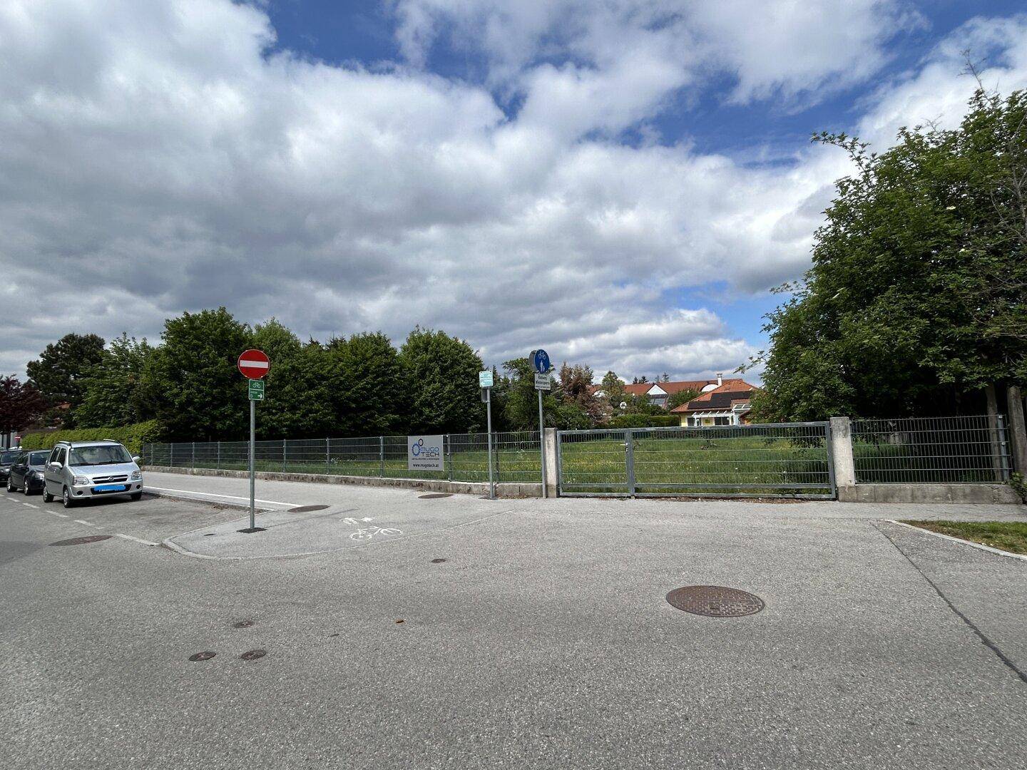 Eine leere Parzelle mit Gras und Bäumen, umgeben von einem Zaun, an einer Straße gelegen.