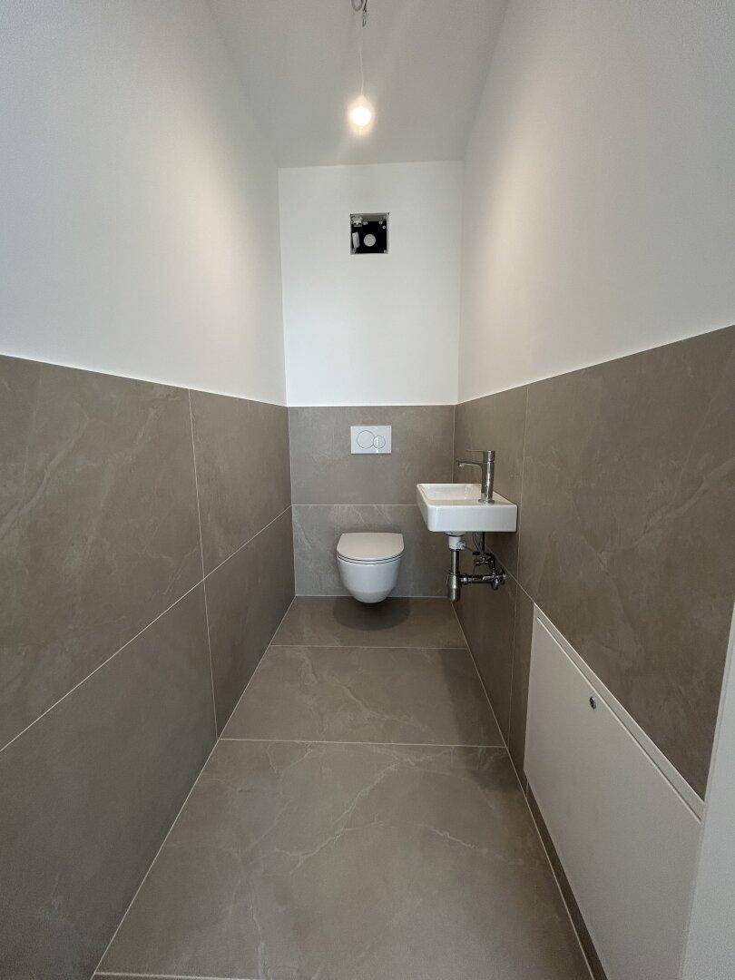 Gäste-WC mit Wand-WC, kleinem Waschbecken und modernen grauen Fliesen.