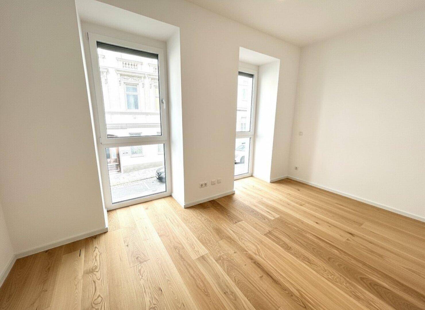 Helles Zimmer mit Holzboden und hohen Fenstern, die einen Blick auf die Umgebung ermöglichen.