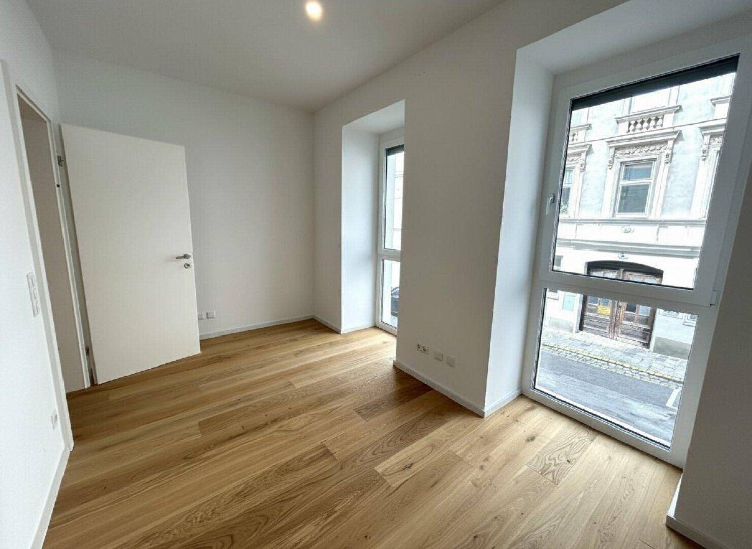 Helles Schlafzimmer mit Holzboden und hohen Fenstern, die einen Blick auf die Straße bieten.