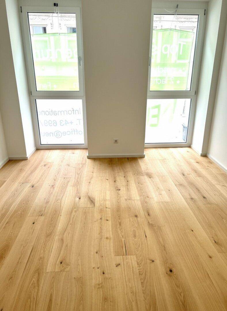 Zimmer mit hellem Parkettboden und zwei hohen Fenstern, die den Raum gut belichten.