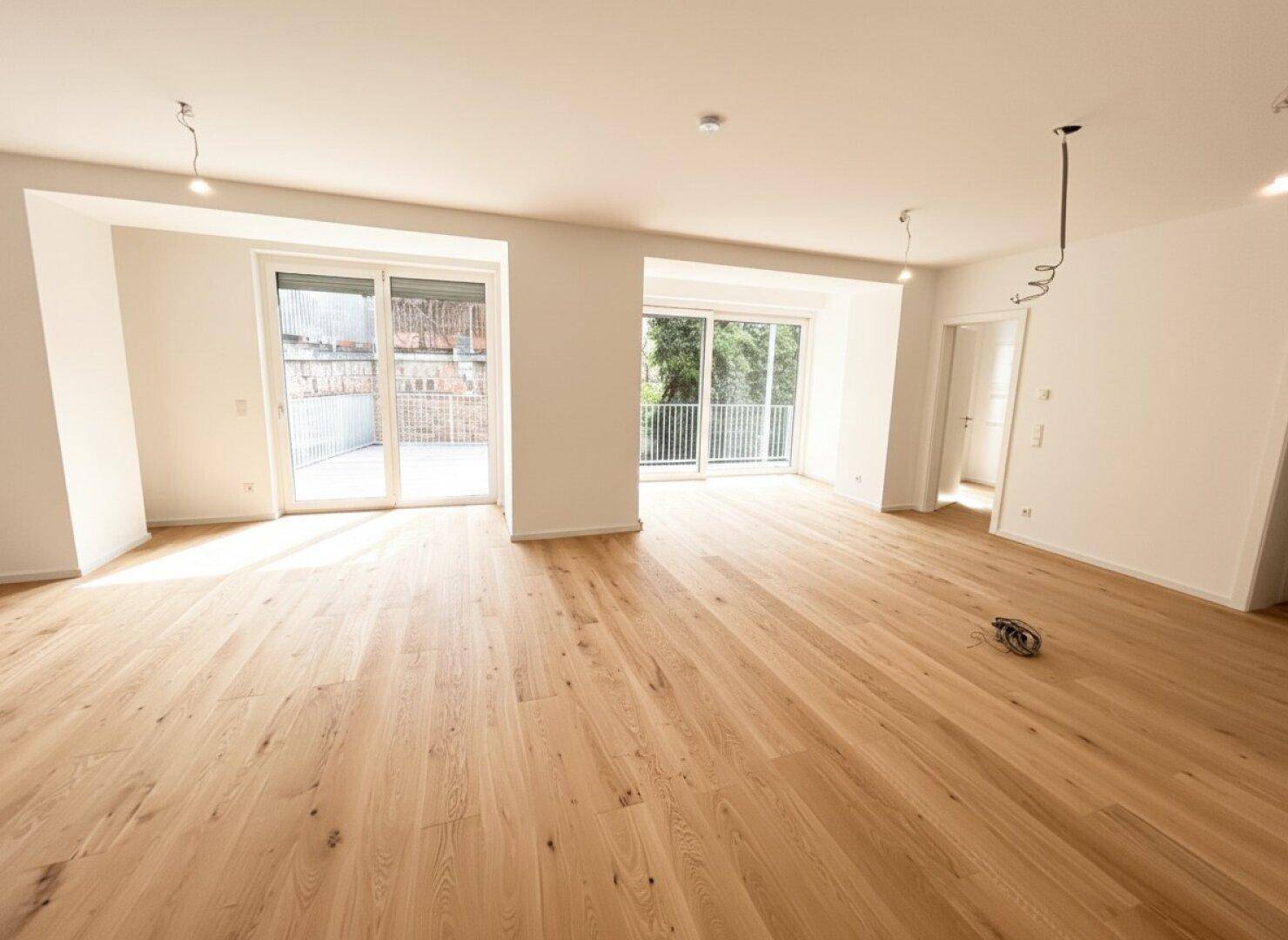 Geräumiges Wohnzimmer mit hellem Parkettboden und großen Fenstern, die viel Tageslicht hereinlassen.