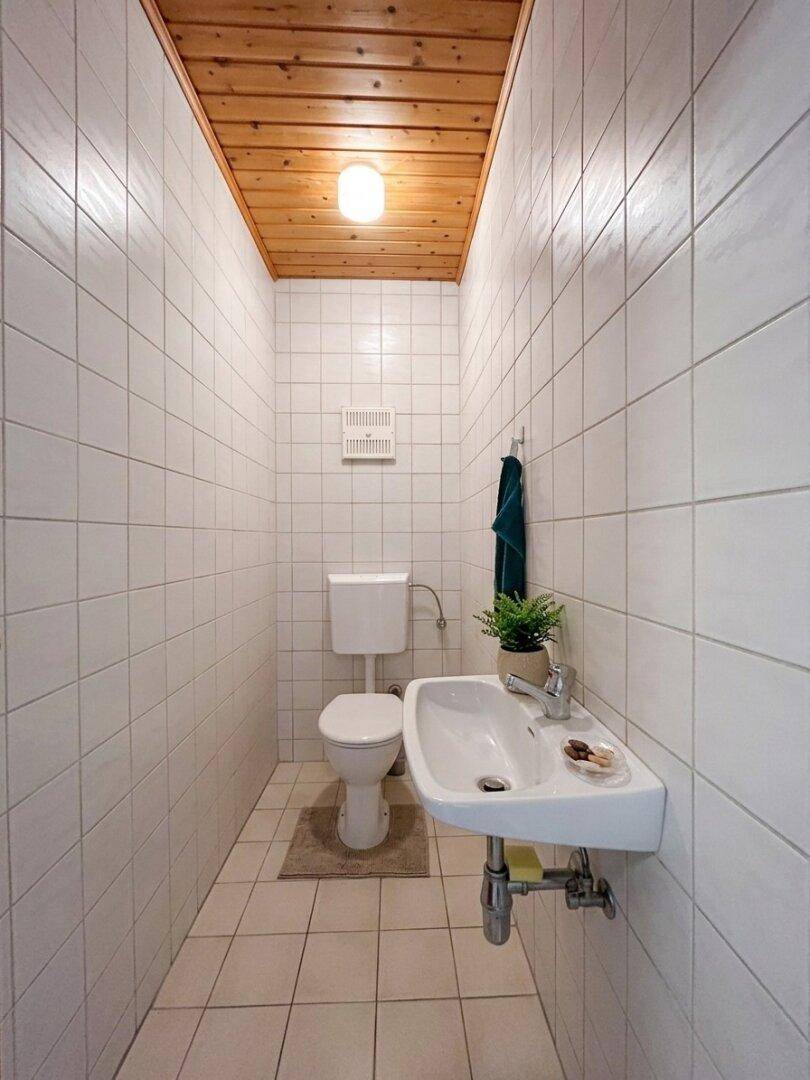 Gefliestes Gäste-WC mit Holzdecke, Toilette, kleinem Waschbecken und Abluftventilator.