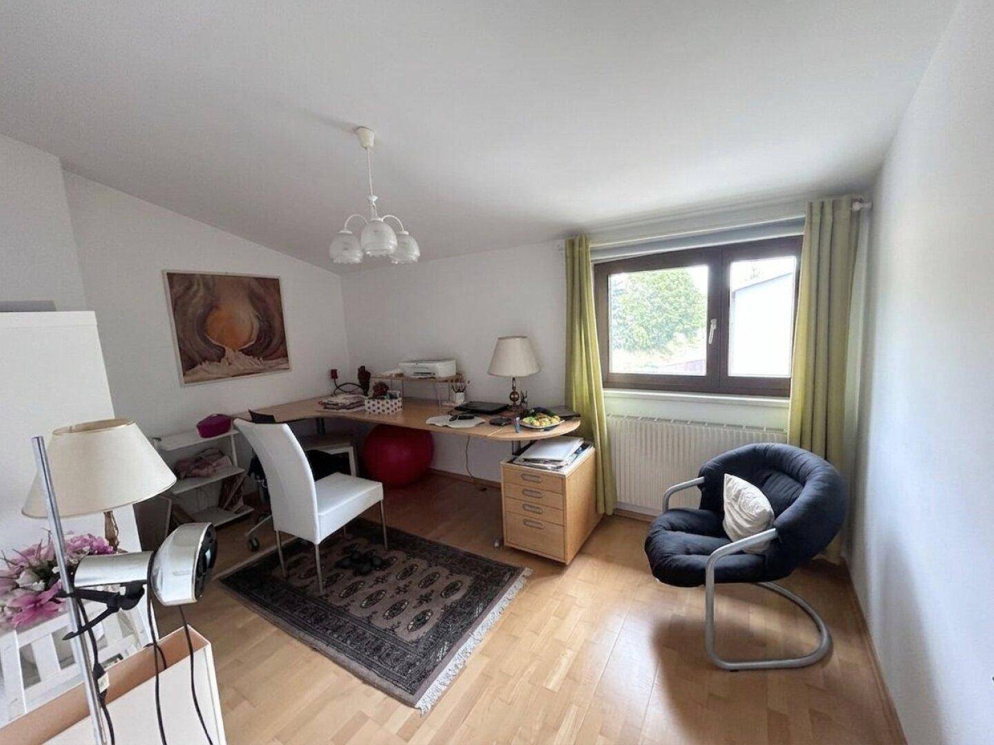 Helles Arbeitszimmer mit Schreibtisch, Bürostuhl und einem bequemen Sessel am Fenster.