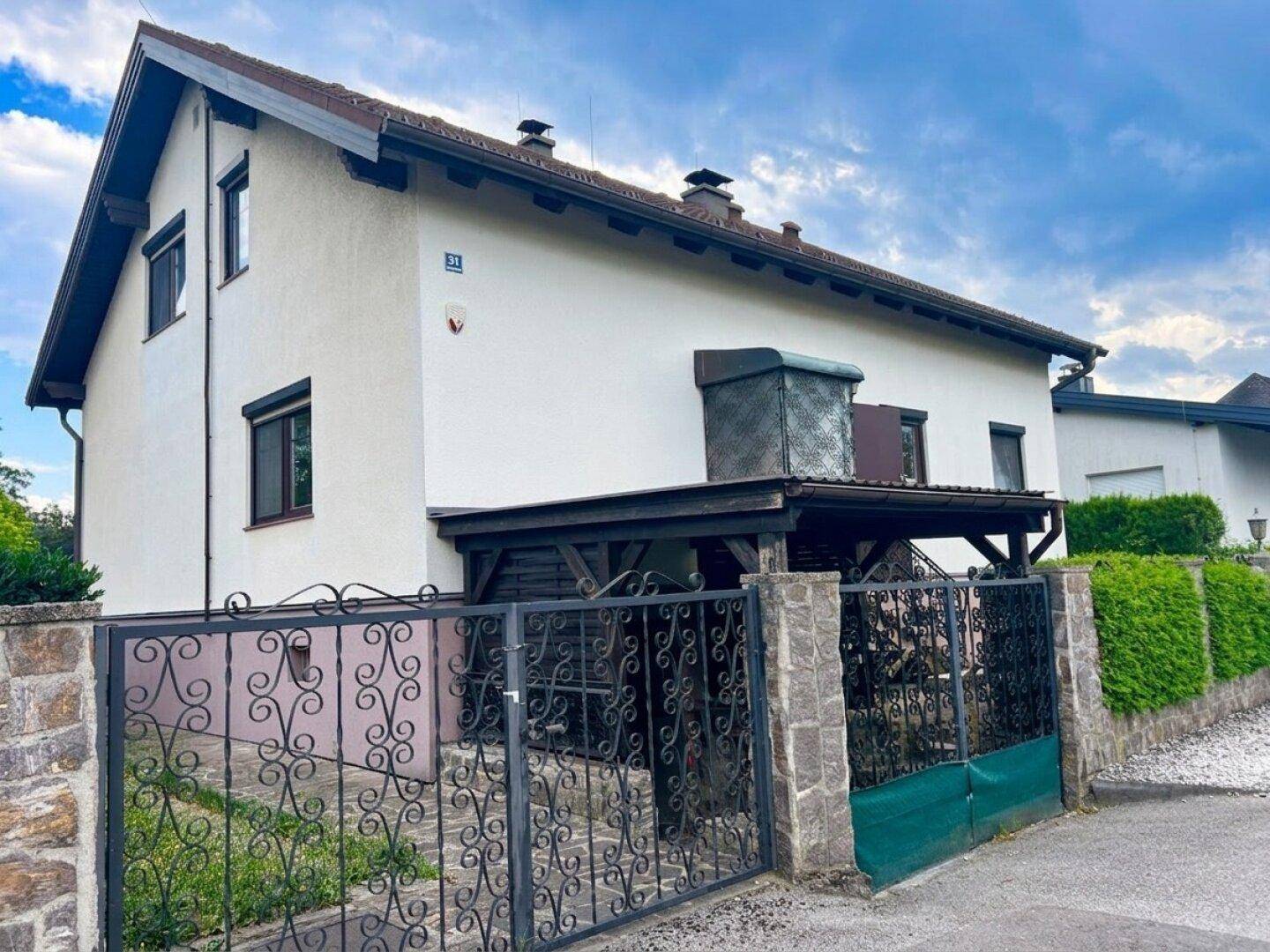 Seitenansicht des Hauses mit überdachtem Eingangsbereich und schmiedeeisernem Tor, umgeben von Grün.