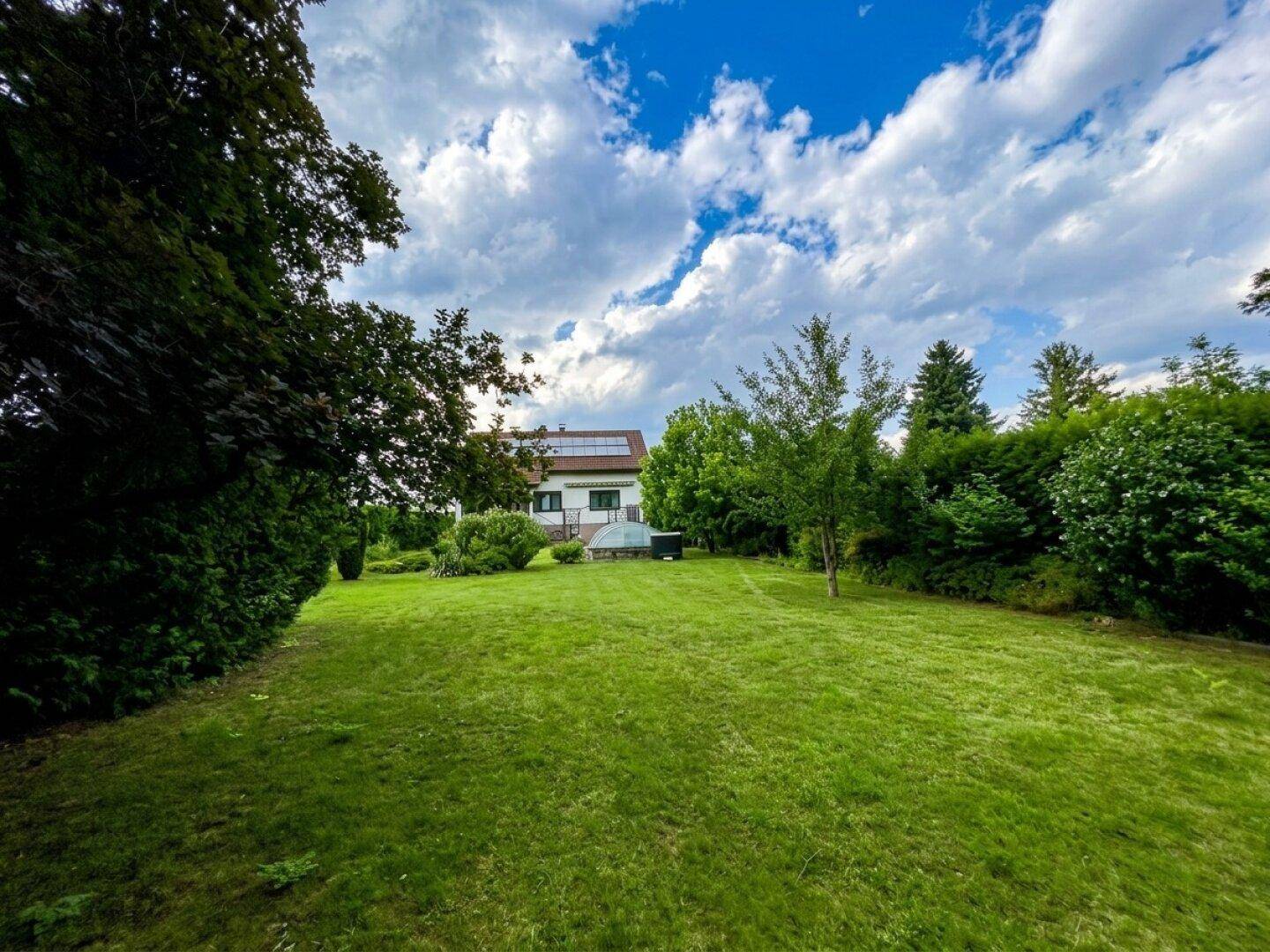 Weitläufige Rasenfläche im Garten mit Bäumen und dem Haus im Hintergrund unter blauem Himmel.