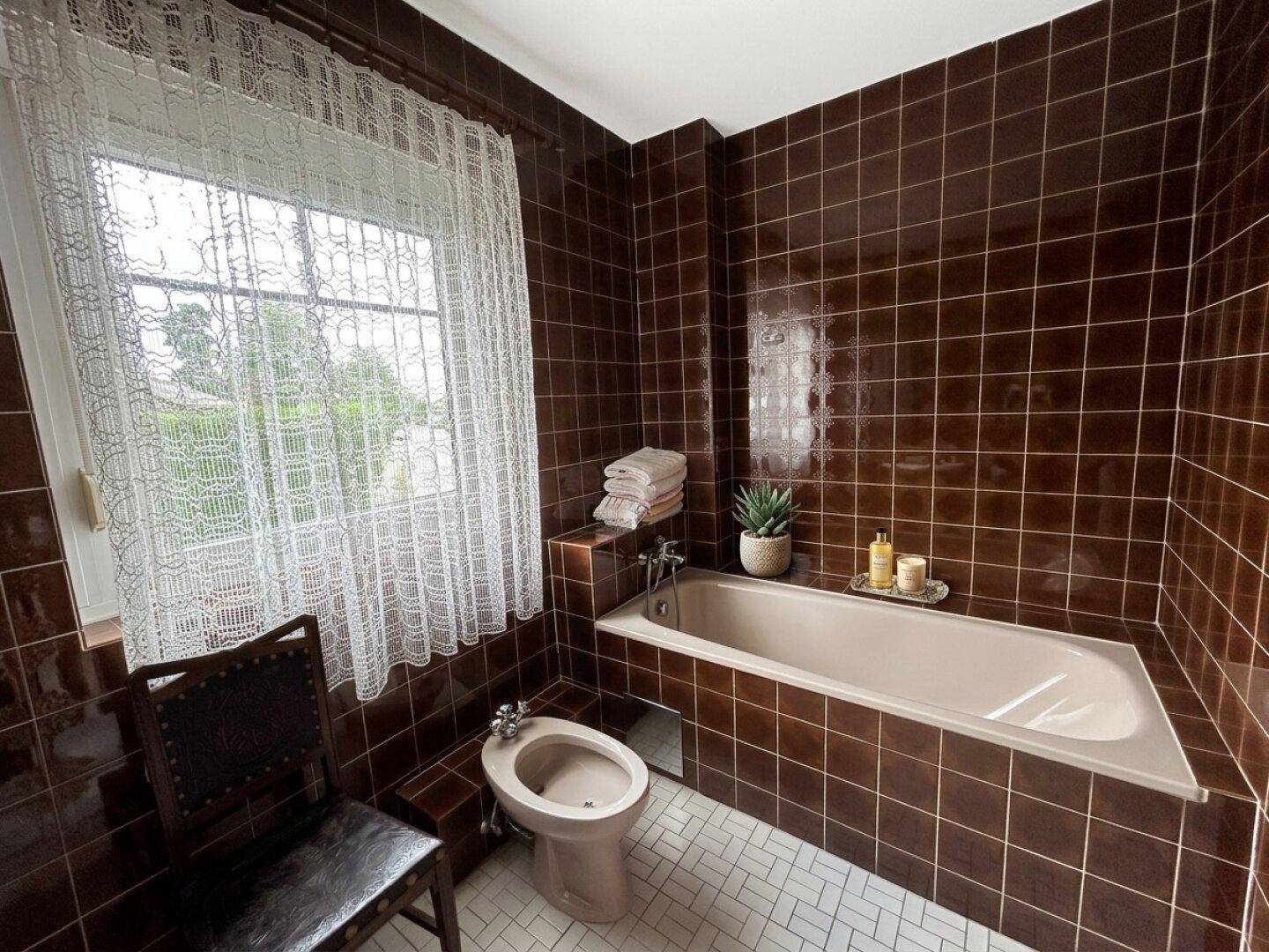 Badezimmer mit braunen Fliesen, Badewanne, Bidet und einem Fenster für Tageslicht.